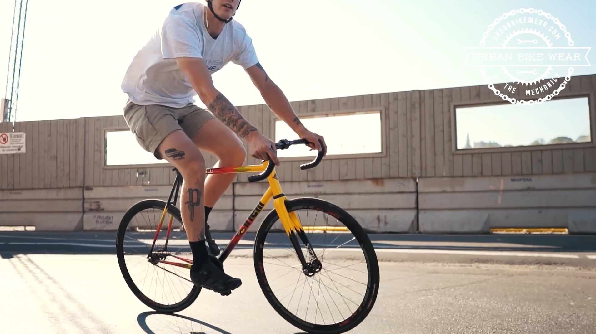 Load video: A young man is riding a brakeless fixed gear bicycle from the brand Cinelli through the streets of sunny Stockholm, Sweden. He is wearing a white T-shirt and khaki shorts. He is also wearing a cycling helmet and white headphones.